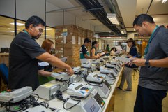 Em primeiro plano, há uma longa bancada com várias urnas eletrônicas dispostas lado a lado, em f...